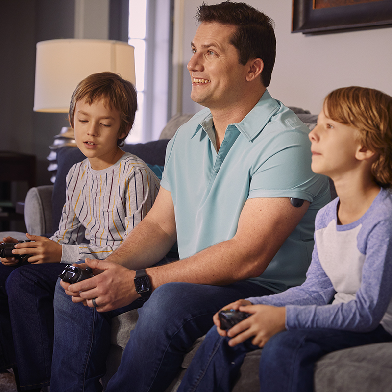 Mann mit implantierbarem CGM am Arm spielt Videospiel mit seinen Kindern